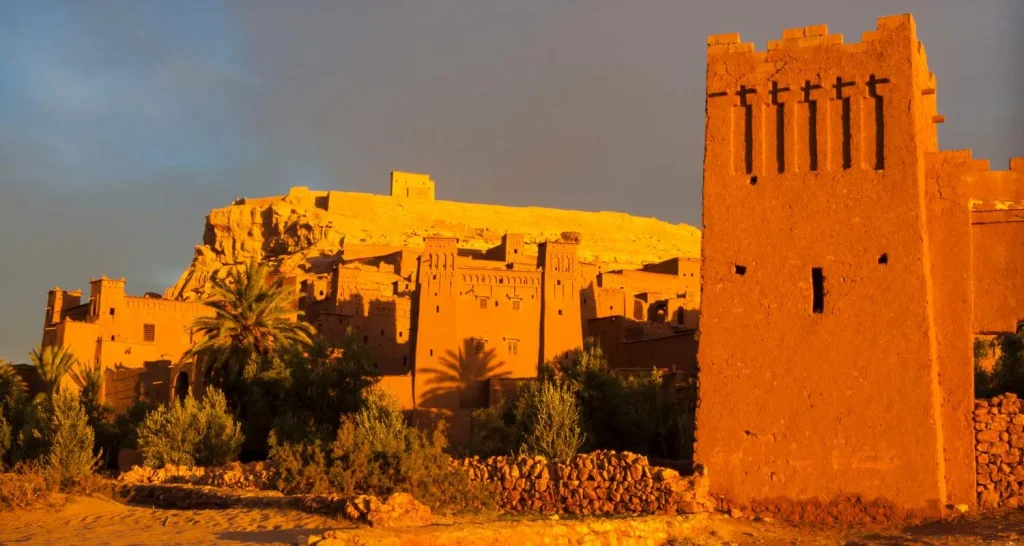 Aït Benhaddou leuchtet im goldenen Licht des Sonnenuntergangs mit einem Wehrturm und einer Palme im Vordergrund